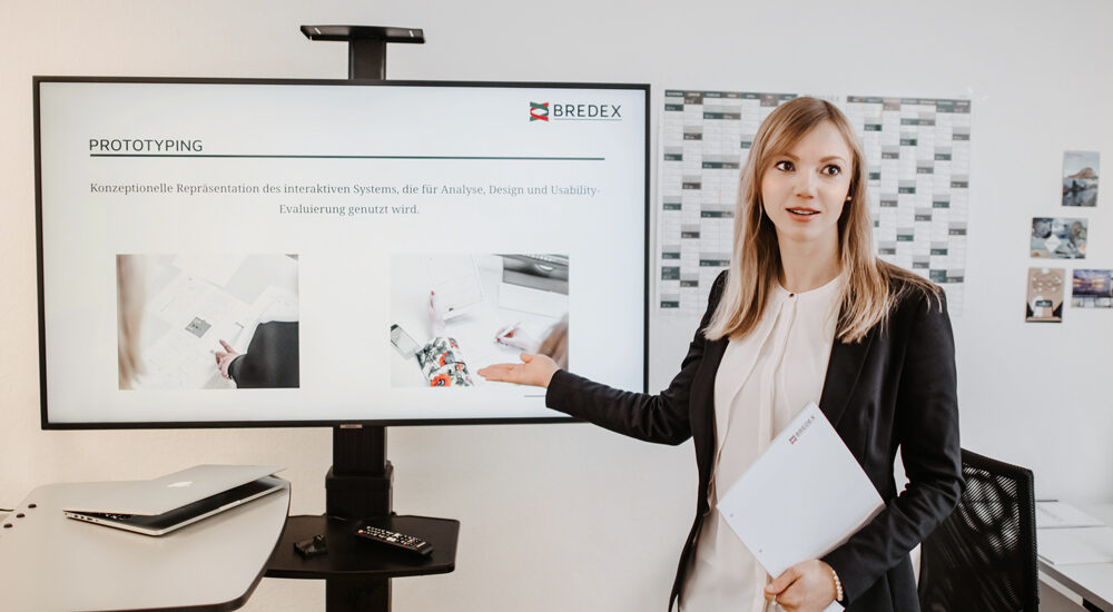 Magdalena Maciejko, UX/UI-Expertin bei BREDEX, steht vor einem Monitor auf dem eine Seite zum Thema Prototyping abgebildet ist.