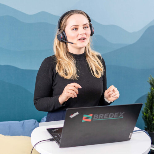 Trainerin Pia Krone, Learning & Development Specialist bei BREDEX, steht mit einem Headset vor ihrem Computer und einer Kamera. Sie gibt eine Remote Schulung im Rahmen des BREDEX Academy Programms.