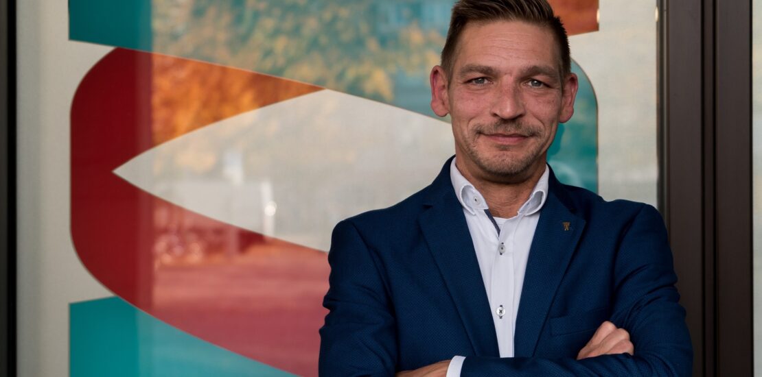 Ron Kneffel, Head of Data Security bei BREDEX, ein Mann mittleren Alters mit kurzen, braunen, zur rechten Seite gekämmten Haaren. Er trägt einen kurzen 3-Tage-Bart. Ron trägt ein dunkelblaues Sakko mit einem weißen Hemd. Die Arme sind vor dem Oberkörper verschränkt.