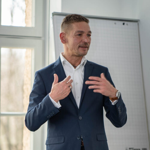 Ron Kneffel, Head of IT Security bei BREDEX, ein Mann mittleren Alters mit kurzen, braunen, zur linken Seite gekämmten Haaren. Er hat ein rundes Gesicht mit etwas weiter auseinander stehenden dunklen Auge. Seine dünnen Lippen sind zu einem leichten Lächeln geformt. Er trägt einen kurzen 3-Tage-Bart. Ron trägt ein dunkelblaues Sakko mit einem weißen Hemd. Er steht vor einer Gruppe von Personen und macht eine erklärende Geste.