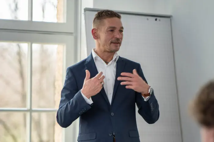 Ron Kneffel, Head of IT Security bei BREDEX, ein Mann mittleren Alters mit kurzen, braunen, zur linken Seite gekämmten Haaren. Er hat ein rundes Gesicht mit etwas weiter auseinander stehenden dunklen Auge. Seine dünnen Lippen sind zu einem leichten Lächeln geformt. Er trägt einen kurzen 3-Tage-Bart. Ron trägt ein dunkelblaues Sakko mit einem weißen Hemd. Er steht vor einer Gruppe von Personen und macht eine erklärende Geste.