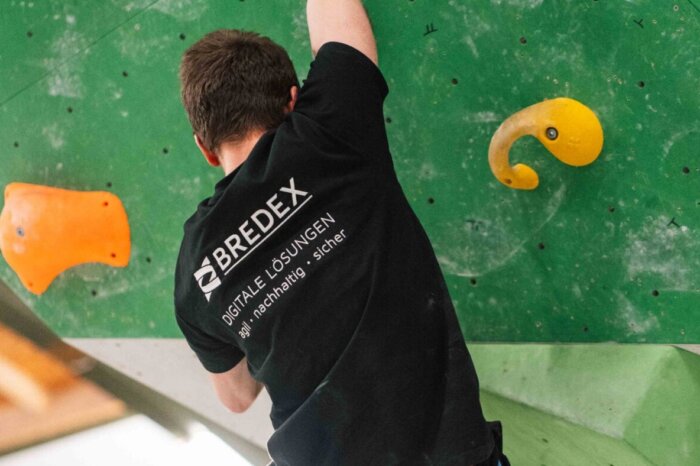 Eine Person beim Bouldern an einer Boulderwand beim Boulder Day. Die Person trägt ein schwarzes T-Shirt mit dem BREDEX Logo.