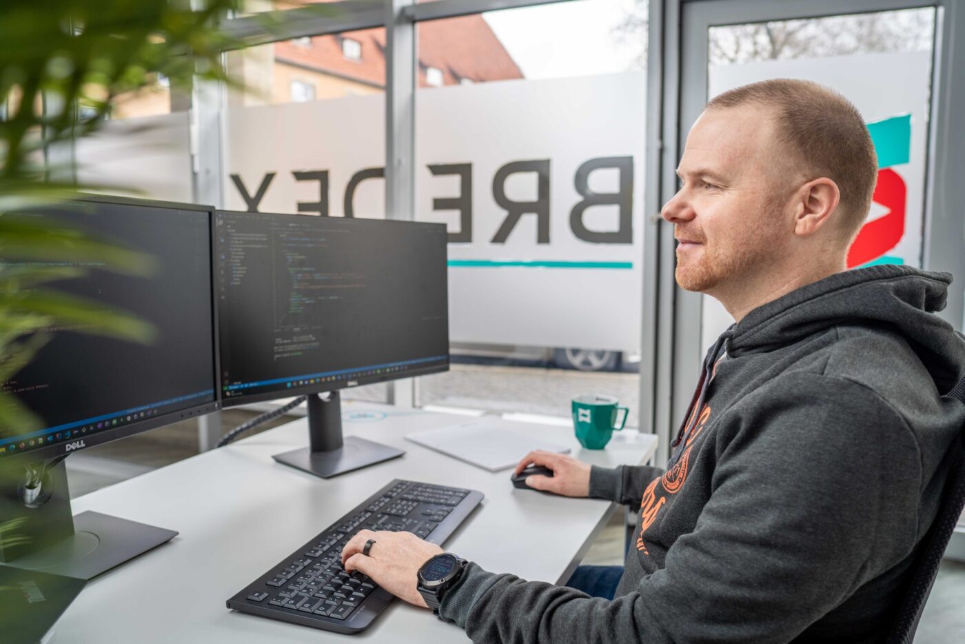 Ein weißer Mann, Softwareentwickler, mit kurzem braunen Haaren und einem Bart sitzt an einem Schreibtisch vor zwei Monitoren. Auf dem Bildschirm sind Codezeilen zu sehen.