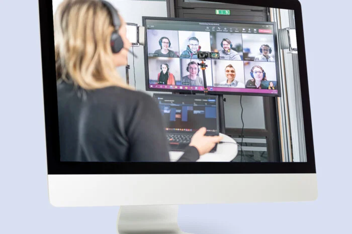 Trainerin Pia Krone steht mit einem Headset vor ihrem Computer und einer Kamera. Sie gibt eine Remote Schulung im Rahmen des BREDEX Academy Programms. Auf einem Monitor dargestellt.