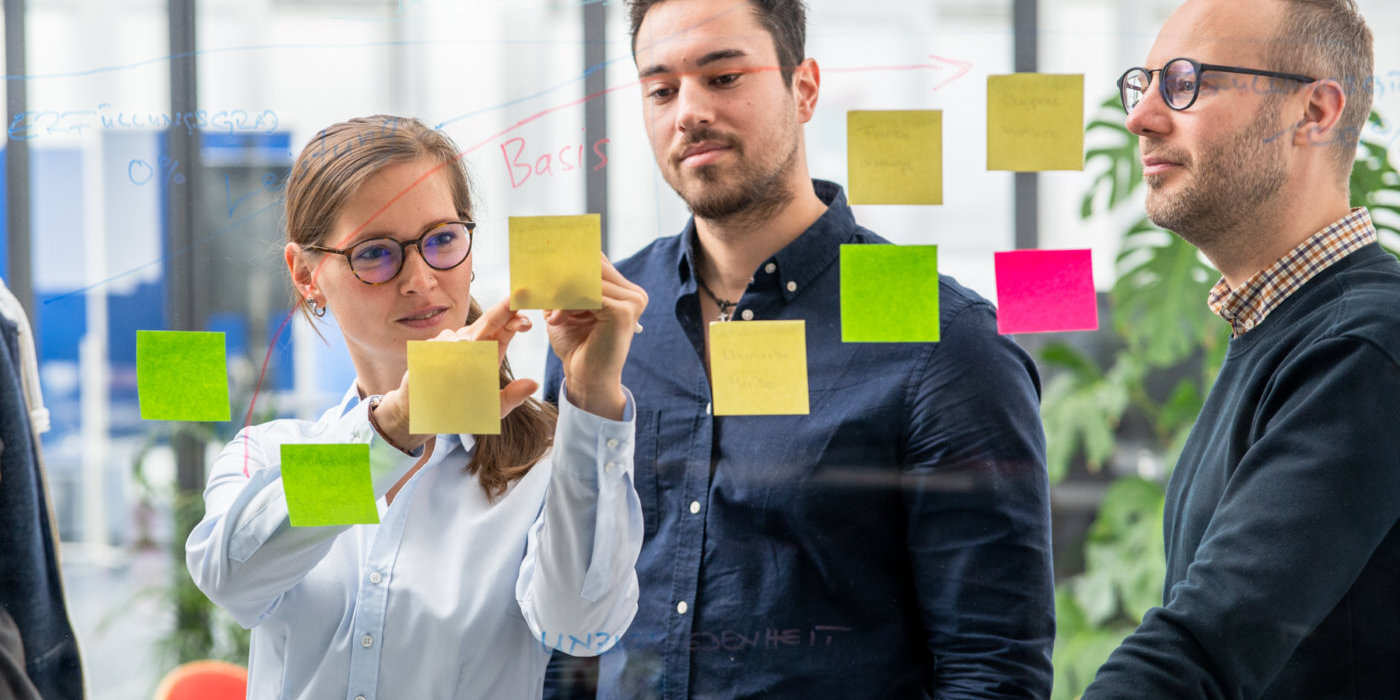 Drei Personen stehen vor einem transparenten Whiteboard. An dem Whiteboard sind mehrere farbige Post-Its befestigt. Diese sind an farbigen Zeichnungen angebracht. Eine weibliche Person beschriftet eines dieser Post-Its, während zwei männliche Personen neben ihr stehen und auf die Beschriftung schauen.