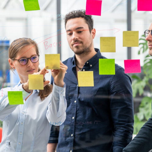 Drei Personen stehen vor einem transparenten Whiteboard. An dem Whiteboard sind mehrere farbige Post-Its befestigt. Diese sind an farbigen Zeichnungen angebracht. Eine weibliche Person beschriftet eines dieser Post-Its, während zwei männliche Personen neben ihr stehen und auf die Beschriftung schauen.
