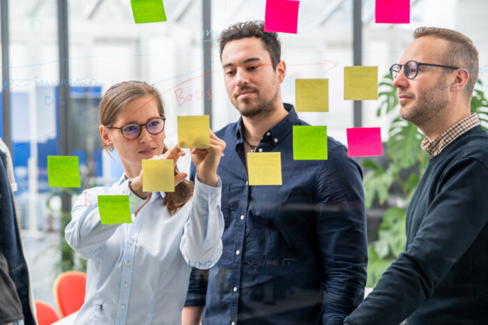 Drei Personen stehen vor einem transparenten Whiteboard. An dem Whiteboard sind mehrere farbige Post-Its befestigt. Diese sind an farbigen Zeichnungen angebracht. Eine weibliche Person beschriftet eines dieser Post-Its, während zwei männliche Personen neben ihr stehen und auf die Beschriftung schauen.