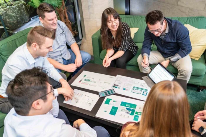 Eine Gruppe von Personen sitzt um einen Tisch herum und sieht sich verschiedenen Arbeitsblätter an, die auf diesem verteilt liegen.