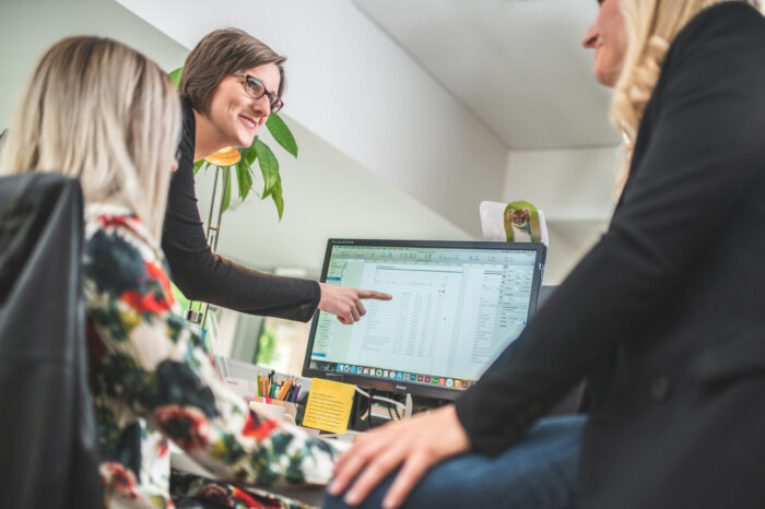 Eine Gruppe von weiblichen Personen, die um einen Monitor herum stehen. Eine der Personen zeigt auf eine Tabelle, die auf dem Monitor abgebildet ist.